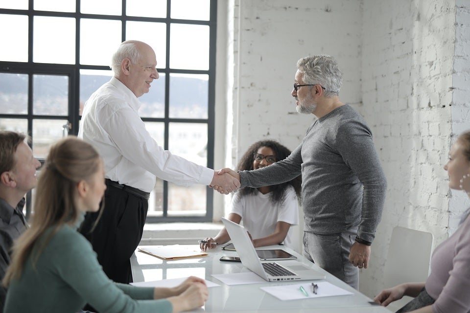 Two men shake hands at a table with a diverse team of employees